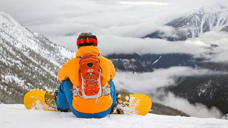 snowboarder in yelow jacket and blue pants sits on snow overlooking the mountains